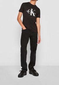 Persona vistiendo una camiseta negra con el logo de Calvin Klein, jeans negros y zapatos de cordones negros, de pie con una mano en el bolsillo.