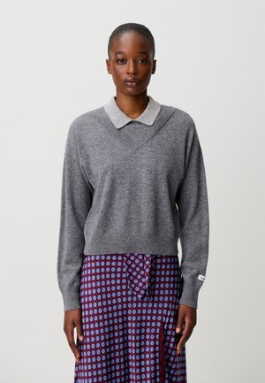 Donna che indossa un maglione grigio sopra una camicia grigio chiaro con colletto e una gonna viola con motivi circolari blu, in piedi davanti a uno sfondo semplice.