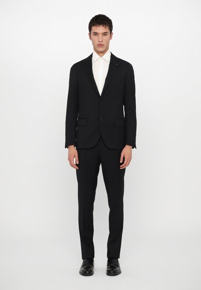 Traje negro con un blazer de un solo botón, dos botones, solapas muescas y pantalones de frente plano. Combinado con una camisa blanca. Zapatos negros.