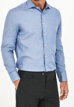 Homme portant une chemise bleu clair à manches longues boutonnée rentrée dans un pantalon noir sur un fond blanc uni.