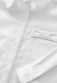 Primo piano di una camicia bianca con rifiniture decorative, bottoni, taschino anteriore e piccolo fiocco di tessuto cucito sulla tasca.