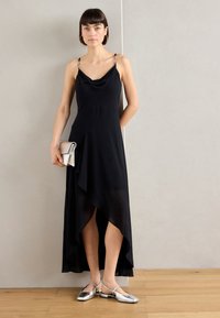 Black asymmetrical dress with a cowl neckline and decorative straps, paired with silver slip-on shoes and a metallic clutch.