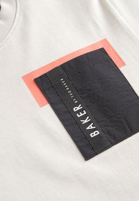 Helebeeži T-särk, millel on silmapaistev must rinnatasku valge logoga ja korallikarva ristkülikukujuline detail tasku kohal. Sile tekstuur.