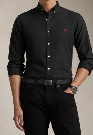Homme portant une chemise noire à boutons avec un logo rouge, un pantalon noir, une ceinture noire et une montre noire au poignet, debout avec une main dans la poche.