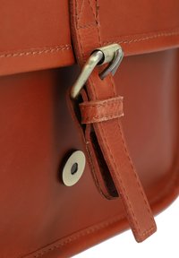 Bruine lederen tas met een gladde textuur, voorzien van een metalen sluiting, een hengsel met stiksel-details en een ronde metalen drukknoopsluiting.