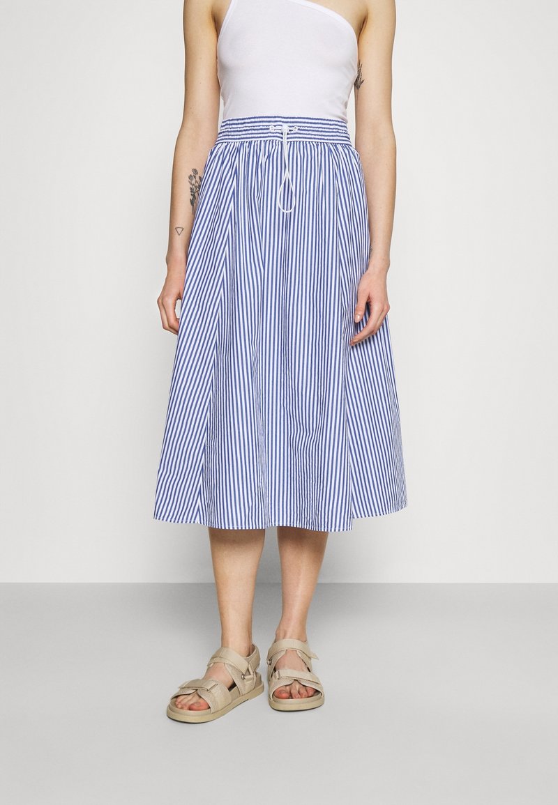 Femme portant une jupe midi rayée verticale bleu et blanc, un top blanc une épaule et des sandales beiges, debout sur un sol gris avec un fond uni.