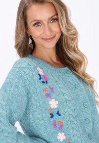 Cardigan de punto en color turquesa con textura de cable, decorado con flores bordadas en rosa, morado y azul. Seis botones azules en la parte delantera.