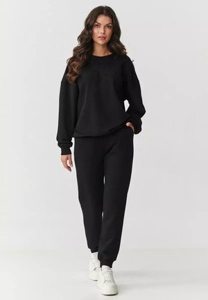 Czarna bluza z długimi rękawami o luźnym kroju, zestawiona z czarnymi spodniami dresowymi. Gładki materiał, minimalistyczny design i białe sneakersy.