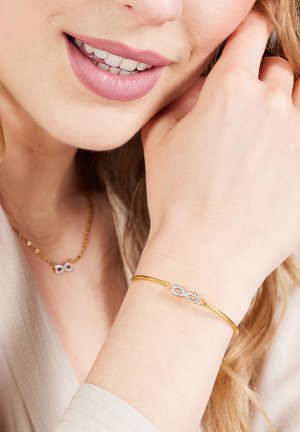 Goldenes Armband mit einem Unendlichkeitssymbol, das mit kleinen klaren Edelsteinen verziert ist. Es wird am Handgelenk getragen, während eine schlichte Kettenkette sichtbar ist.