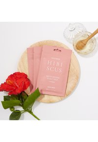 Three pink pouches labeled "HIBISCUS Moisturizing Scalp Pack" on a wooden plate, with a red hibiscus flower and a glass jar of honey.