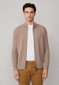 Beżowy kardigan z warkoczowym splotem, z wysokim kołnierzem i zamkiem, z prążkowanymi mankietami, zestawiony z białym T-shirtem i brązowymi chinosami.