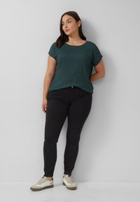Donkergroen structuur t-shirt met korte mouwen, gecombineerd met zwarte slim-fit jeans en witte sneakers met bruine accenten, tegen een eenvoudige achtergrond.