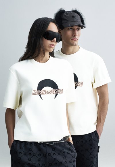 T-shirts en coton blanc avec un grand logo croissant noir et le texte « MARINE SERRE ». Le modèle masculin porte un chapeau noir avec des accents d'oreilles en fourrure.