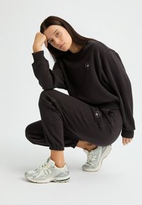 Sudadera negra y pantalones de chándal de tela suave, con un pequeño logo en el pecho y en el bolsillo. Zapatillas gris y blancas con detalles sutiles.