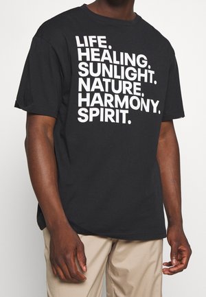 Mężczyzna w czarnej koszulce z białym, pogrubionym tekstem wymieniającym: Życie. Uzdrawianie. Światło słoneczne. Natura. Harmonia. Duch. Widoczne beżowe spodnie.