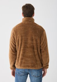 Barna bolyhos dzseki magas gallérral, texturált, puha anyagból, hosszú ujjú, kék farmernadrággal párosítva. Hátsó nézet, minimális részlet.