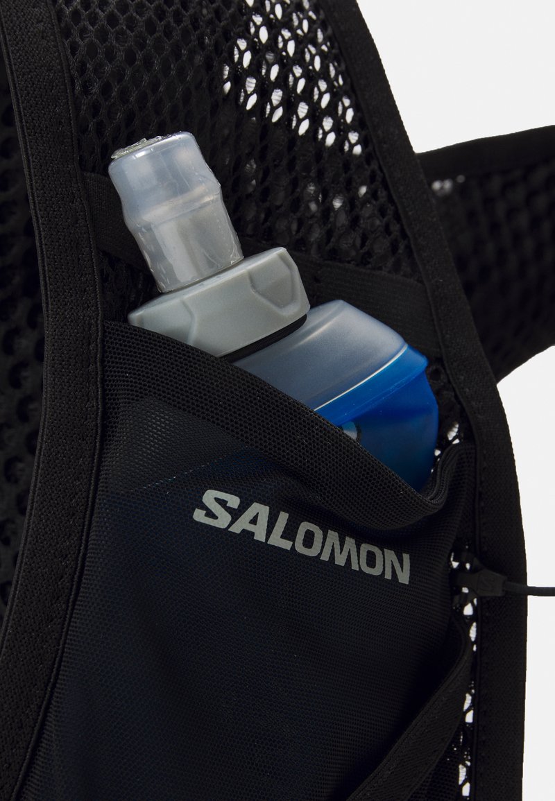 Zaino per idratazione in rete nera con una bottiglia d'acqua blu parzialmente visibile. Presenta il logo grigio Salomon, un design elegante e spallacci regolabili.