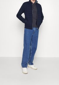 Chaqueta de cremallera acolchada navy sobre una camisa polo oscura, combinada con unos vaqueros de pierna ancha azules y zapatillas blancas, frente a un fondo sencillo.