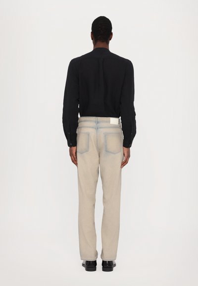 Pantaloni in denim beige con accenti azzurri, tasche posteriori e un design pulito. Indossati con una maglietta nera a maniche lunghe e scarpe nere.