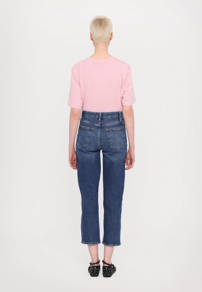 Pink short-sleeve shirt paired with high-waisted blue denim jeans. Jeans feature back pockets and a cropped cut above the ankle. Black studded shoes.