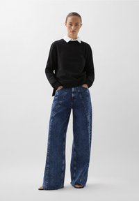 Sudadera negra con cuello blanco, combinada con vaqueros de mezclilla azul de pierna ancha que cuentan con bolsillos frontales y un patrón desteñido.