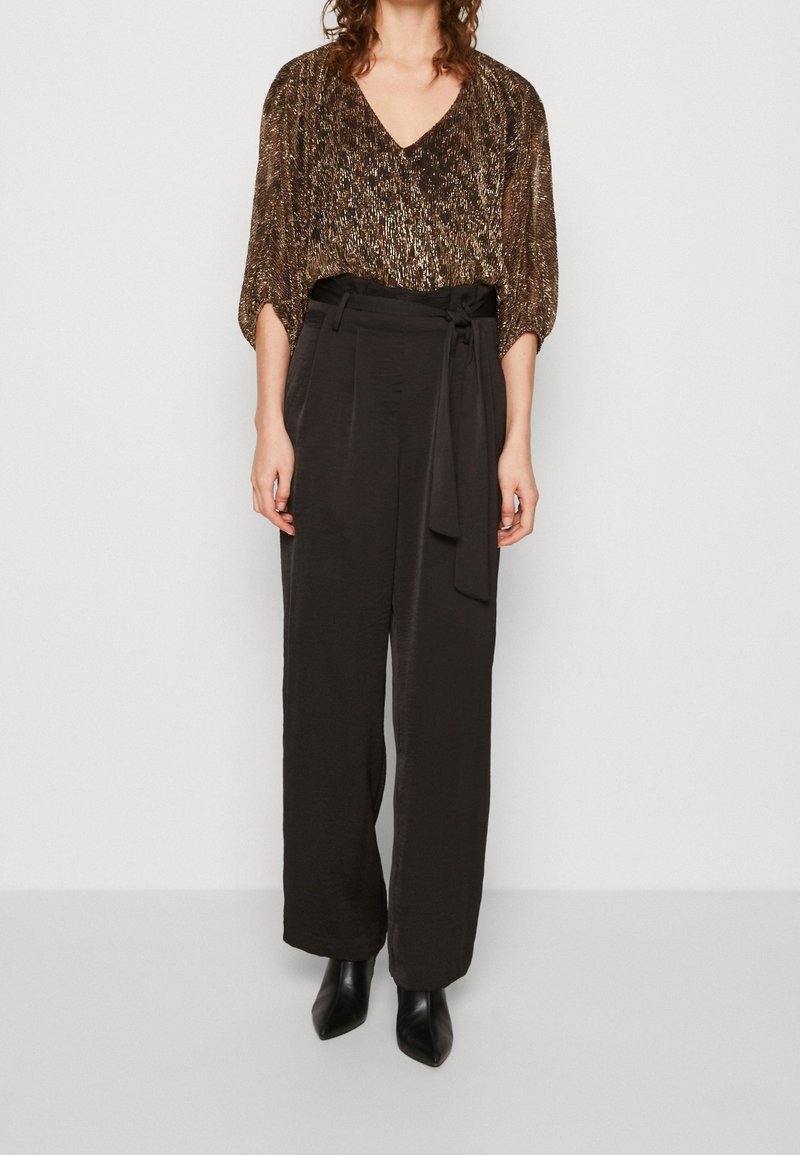 Femme portant un chemisier métallique doré à col en V, un pantalon noir taille haute à jambes larges avec un lien à la taille, et des bottes noires à bout pointu.