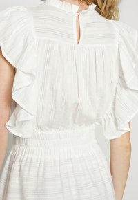 Chemisier blanc avec des manches froncées, un motif rayé texturé et une ouverture en forme de goutte au dos avec un bouton. Détail de taille élastique.