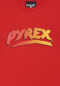 Maglietta in cotone rossa con un grande logo grafico "PYREX" multicolore. Il design presenta uno stile audace con tratteggi di pennello e una sfumatura arancione.