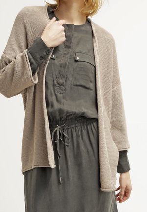 Person wearing a taupe knitted cardigan over a dark gray long-sleeve dress with a drawstring waist and chest pocket with flap.