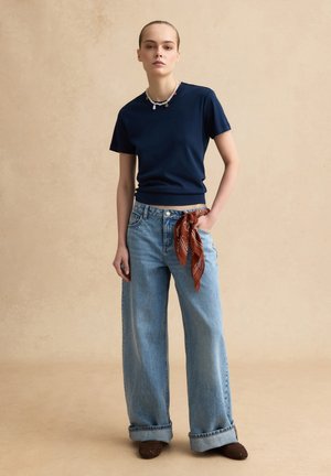 Jonge vrouw staand, gekleed in een marineblauw shirt met korte mouwen, brede lichtblauwe jeans met een bruine sjaal die aan de zak is geknoopt, en bruine schoenen.