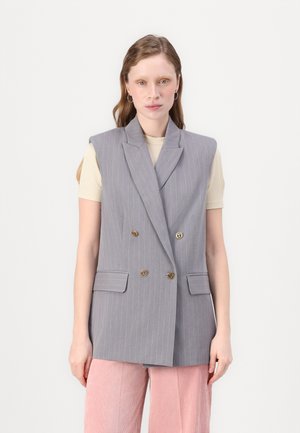 Gilet croisé à rayures grises avec épaulettes, boutons dorés et deux poches avant, associé à un haut beige clair et un pantalon rose.