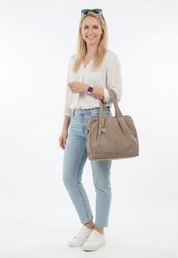 Lächelnde Frau trägt weiße Bluse, hellblaue Jeans, weiße Turnschuhe, lila Uhr, Sonnenbrille auf dem Kopf, hält eine beige Tragetasche vor weißem Hintergrund.
