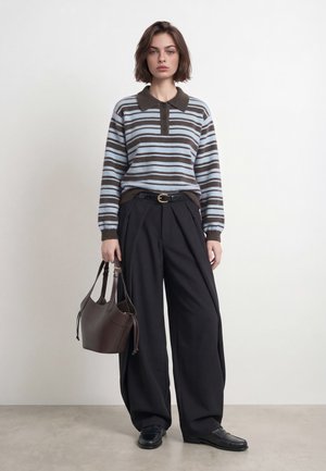 Un mannequin se tient dans un studio, vêtu d’un pull à col rayé bleu et marron, d’un pantalon noir ample et de mocassins, et tient un sac à main marron.