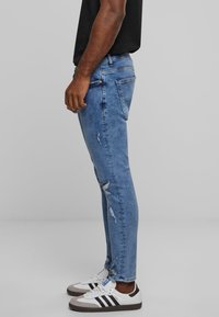 Jeans ajustados azul con un acabado desgastado, que presentan desgastes en la rodilla y cinco bolsillos, combinados con zapatillas blancas y una camiseta negra.
