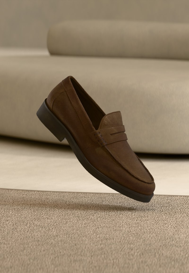 Καφέ μοκασίνι από σουέτ με μαύρη σόλα, αιωρούμενο πάνω από μπεζ χαλί μπροστά από μπεζ καναπέ.