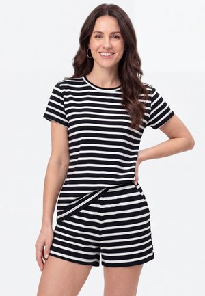 Glimlachende vrouw met lang bruin haar draagt een zwart-wit horizontaal gestreept kortemouwen topje en bijpassende shorts tegen een effen witte achtergrond.