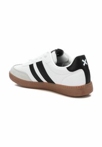Zapatilla deportiva blanca con rayas negras, con un talón negro texturizado y una suela de goma. Lengüeta ligeramente acolchada y diseño de cordones.