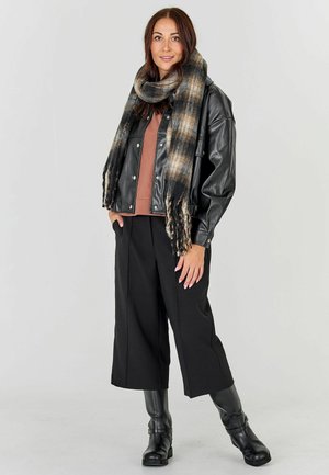 Moteris dėvi juodą odinę striukę, rudą palaidinę, juodus plačialygius kelnes, juodus batus ir didelį languotą šalį, stovinti priešais vienspalvį foną.