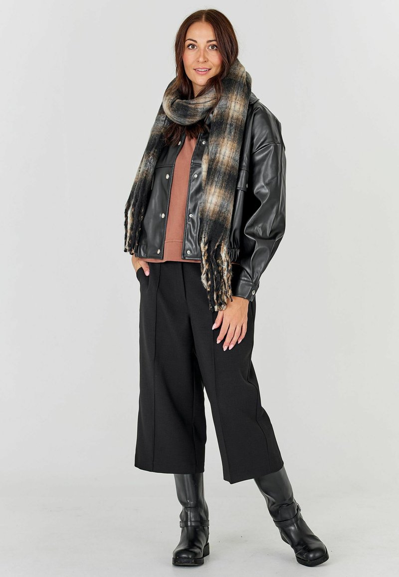 Femme portant une veste en cuir noire, un haut marron, un pantalon large noir, des bottes noires et une grande écharpe à carreaux, debout devant un fond uni.
