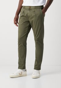 Pantaloni verde măsliniu cu pliseuri, talie elastică și închidere cu nasture. Asortați cu adidași albaștri cu accente bej.