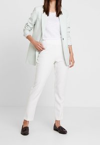 Lichtgroene op maat gemaakte blazer over een witte textuur shirt, gecombineerd met een fitte witte broek en donkere instappers. Blazer heeft opgerolde mouwen.