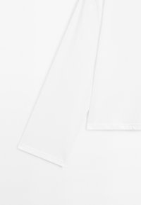 Witte langslokshirt met eenvoudige stiksels op een witte achtergrond, waarbij een deel van de mouwen en de zoom zichtbaar is.