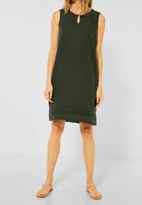 Ärmelloses olivgrünes Kleid mit einem Schlüssellochausschnitt, strukturiertem Stoff und horizontalen Akzentlinien am Saum. Kombiniert mit rosa Flip-Flops.