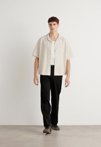 Kortsleeve, skjortebluse i lys beige, paret med svarte, rette bukser. Modellen står foran en ensfarget bakgrunn.