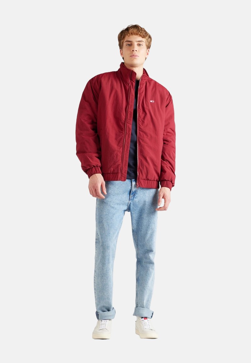 Chaqueta bomber roja con cierre frontal, puños elásticos y un pequeño logo. Combinada con jeans de color azul claro y zapatillas blancas. Diseño simple y casual.