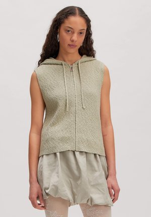 Frau trägt ärmellosen strukturierten Hoodie mit Kordelzügen, lockerem Satinsrock und Strumpfhosen mit Spitzenmuster, steht vor einfarbigem Hintergrund.