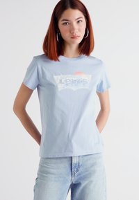 Šviesiai mėlyna medvilninė marškinėliai su balta Levi's logotipo grafika priekyje. Trumpomis rankovėmis ir laisvu siluetu, derinami su šviesiai mėlynomis džinsais.