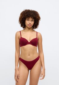 Γυναίκα με σγουρά μαλλιά που φοράει ένα ταιριαστό σετ σουτιέν και εσώρουχου σε σκουροκόκκινο χρώμα, στέκεται μπροστά σε ένα απλό ανοιχτό γκρι φόντο.