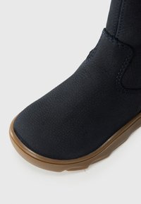 Marineblauwe boot met een gestructureerde synthetische bovenkant, contrasterende bruine zool en subtiele blauwe stiksels langs de naden. Brede teenvorm.