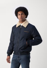Chaqueta de bomber azul marino con un cuello de piel beige, cierre frontal, puños de canalé y un parche de logo en el pecho. Llevada con jeans de mezclilla oscuros.